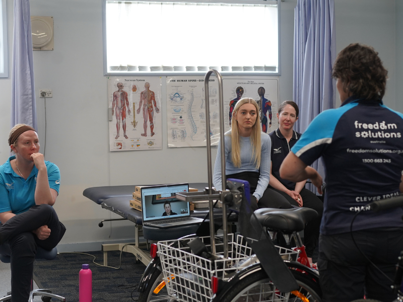 A woman from Freedom Solutions Australia, seen from behind, is speaking to two women seated in a clinical setting. The two women are listening attentively. In the foreground, an adaptive tricycle with a basket is visible. On the wall behind them, anatomical posters depicting the human spine and muscular system are displayed. A laptop to the side shows a person participating in the session virtually. The environment suggests an educational or training session focused on assistive technology and its application in therapy.