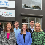 Group of seven people smiling and standing outside the Freedom Solutions Australia building, which displays signs for both 'Freedom Solutions Australia' and 'Freedom Wheels.' The individuals are dressed in business casual attire, with the building's large windows visible behind them.