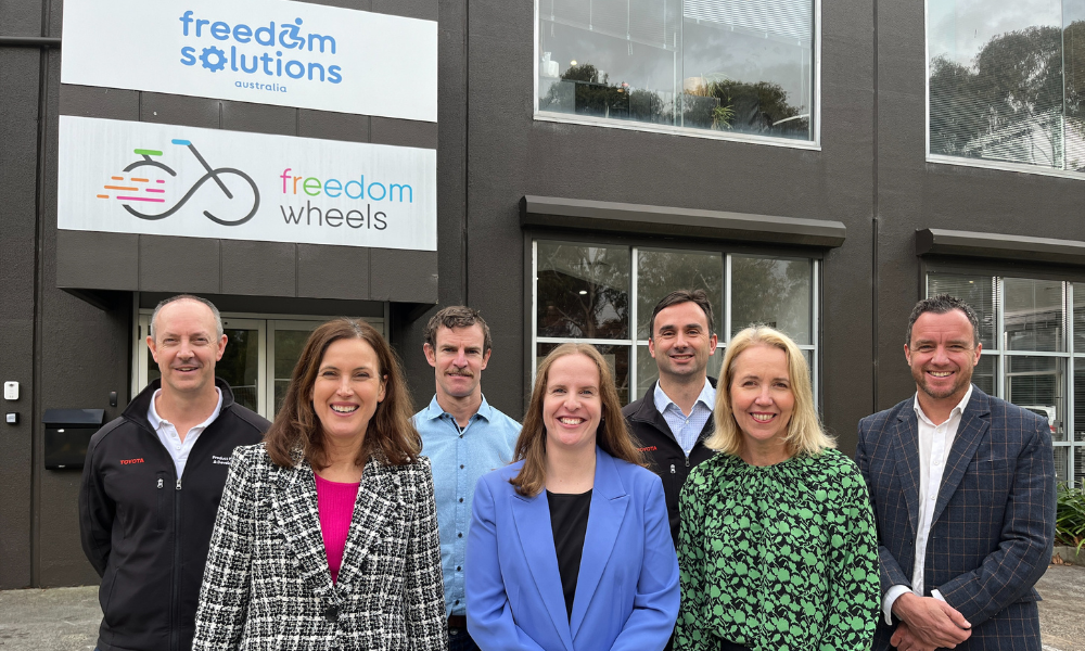 Group of seven people smiling and standing outside the Freedom Solutions Australia building, which displays signs for both 'Freedom Solutions Australia' and 'Freedom Wheels.' The individuals are dressed in business casual attire, with the building's large windows visible behind them.