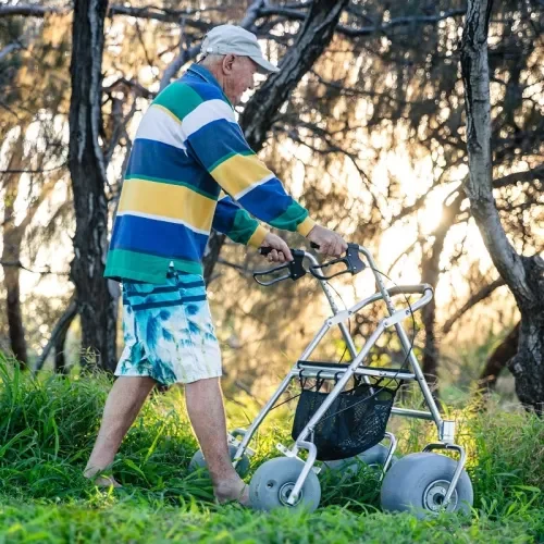 An elderly man is outdoors, walking with the assistance of a sturdy rollator walker. He is dressed casually in a colourful striped shirt, blue and white tie-dye shorts, and a light-coloured cap. The background shows a natural setting with trees and sunlight filtering through, creating a serene atmosphere.