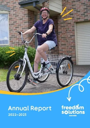 Cover page of the Annual Report 2022–2023 for Freedom Solutions Australia. The top half features a smiling woman wearing a helmet, riding a white tricycle in front of a brick house. Yellow lines around her indicate joy and excitement. The bottom half of the cover is blue with the Freedom Solutions Australia logo on the right, and the text "Annual Report 2022–2023" on the left. A white arrow with a curly line points from the tricycle to the logo.