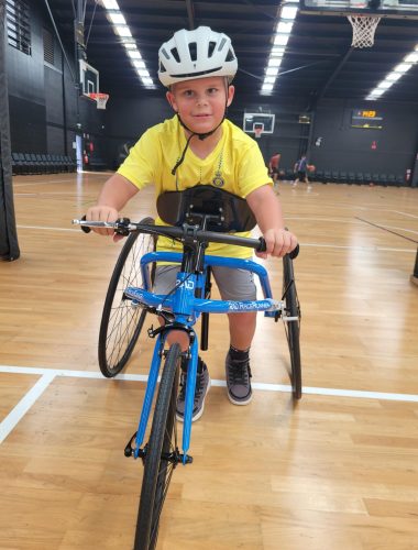 A young boy wearing a yellow shirt and a white helmet is riding a blue adaptive tricycle indoors. He is in a large gymnasium with basketball hoops and wooden flooring. The boy is looking directly at the camera and smiling.