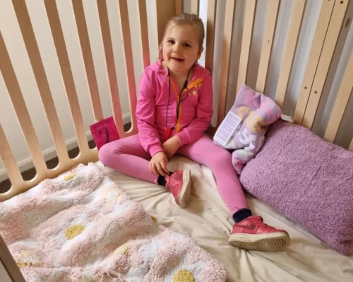 The image depicts a young girl sitting in a cozy, safe, and secure enclosed bed. She is wearing a pink outfit, matching the pink and white bedding and pillows around her. The girl appears happy and comfortable, highlighting a supportive environment. The bed's design, with its protective wooden slats, suggests it is tailored for children who need a secure sleeping space due to health or safety concerns. This setup illustrates the importance of customised sleeping solutions in ensuring the well-being and comfort of children with special needs.