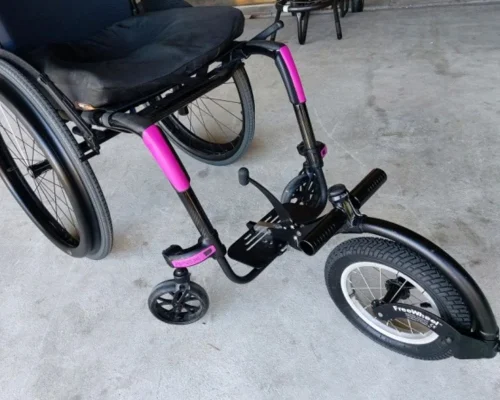 The image shows a wheelchair with a specialized front wheel attachment designed for improved mobility and maneuverability. The attachment features a large front wheel, similar to a bicycle wheel, which likely helps in navigating uneven surfaces more smoothly. The frame of the wheelchair and the attachment includes black and vibrant pink elements, giving it a stylish appearance. The wheelchair itself appears to be a manual model with a cushioned seat for comfort. This adaptive equipment likely enhances the user's independence and ability to traverse various terrains.