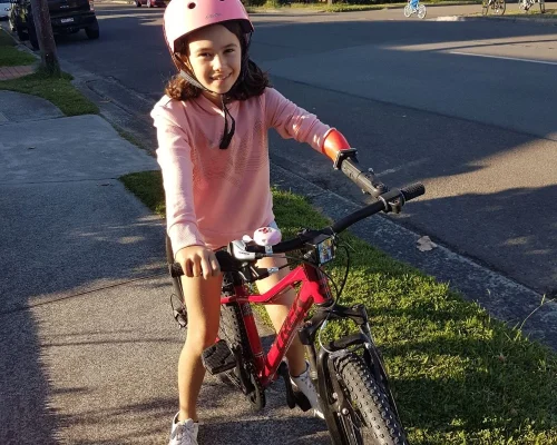 The image shows a young girl wearing a pink helmet and a matching pink long-sleeved shirt, riding a red bicycle on a suburban street. She is smiling at the camera and appears to be enjoying her time. Her right arm has a bright orange prosthetic, which is designed to help her grip and control the bike's handlebars. The background includes a few other children riding their bikes and cars parked along the street, suggesting a typical, safe neighbourhood setting. The overall atmosphere of the image is positive and highlights the girl's independence and confidence while riding her bike