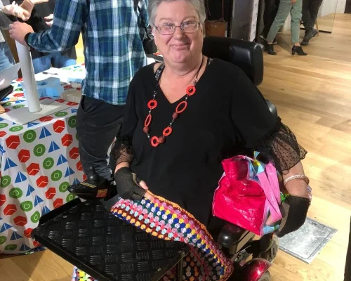 The image shows a woman in a motorised wheelchair, smiling warmly at the camera. She is wearing a black outfit with a red and black necklace and has a colourful crocheted blanket over her lap. The woman is in an indoor setting that appears to be a community event or gathering, as evidenced by people interacting in the background and a table covered with a brightly patterned tablecloth. The environment looks lively and inclusive, suggesting a supportive community atmosphere.