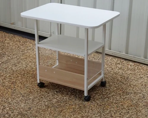 The image depicts a small, white utility table with a minimalist design. The table has a rectangular top surface and an additional shelf below. It is supported by a metal frame with four legs, each equipped with a caster wheel for mobility. The bottom section features a wooden panel that likely serves as a base or storage compartment. The table is positioned on a textured outdoor surface with a metal panel background, indicating it might be used in a versatile setting, both indoors and outdoors.