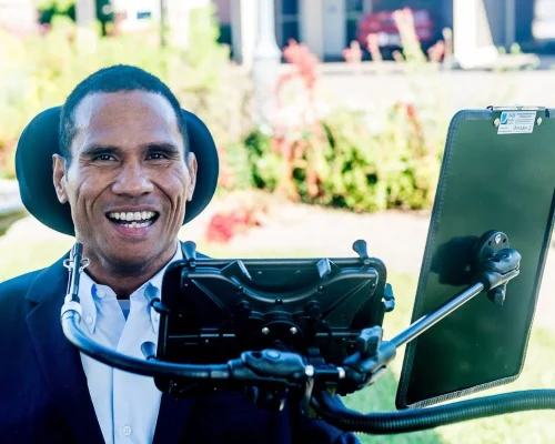 In an outdoor setting, a person is sitting in a wheelchair, equipped with assistive technology. The individual, dressed in a dark suit jacket and a light blue shirt, is smiling broadly, exuding a positive and joyful demeanor. The wheelchair is equipped with a mounted device, likely a communication aid or tablet, which is positioned in front of them. The background features greenery and a residential area, suggesting a pleasant and inclusive environment.