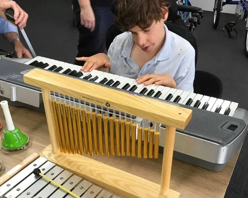 In an indoor setting, a young person with brown hair is engaged in playing a keyboard. They are seated in a wheelchair and seem to be deeply focused on their music. In the foreground, there is a wooden percussion instrument with metal chimes and a xylophone, indicating a rich musical environment. Other people are present in the background, suggesting a supportive and inclusive setting where the young person is encouraged to explore and enjoy music.