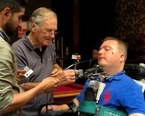 "In an indoor setting, a man in a blue polo shirt is seated in a wheelchair, receiving assistance from two other men. The man in the wheelchair has various assistive devices attached to his chair and body, aiding his communication and mobility. One man, who appears to be elderly and wearing glasses, is carefully adjusting a device while engaging in conversation. The other man, possibly a technician or volunteer, is also focused on the device. The scene depicts a collaborative effort to enhance the quality of life and independence for the man in the wheelchair."