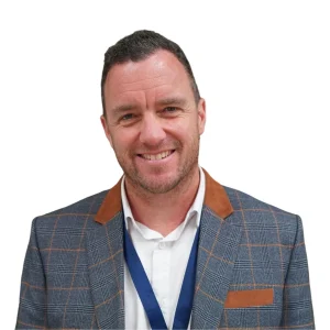 A professional headshot of a middle-aged man with short brown hair, wearing a grey checked blazer with brown accents and a white shirt. He is smiling and looking directly at the camera. The background is plain white.