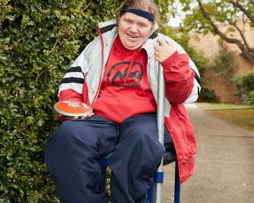 In a bright and cheerful outdoor setting, a woman with a broad smile is sitting comfortably on a chair. She is dressed warmly in a red hoodie with 'NRG 2019' printed on it, a grey and white jacket, and dark pants. She has a headband keeping her hair back. In one hand, she holds an orange discus, while her other hand is gripping a supportive pole. The background features lush green foliage, adding to the vibrant and positive atmosphere of the scene.