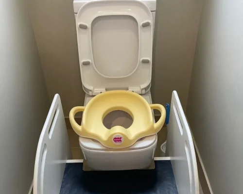 In a small bathroom, there is a white toilet equipped with a yellow potty training seat. The seat has two large handles for stability and ease of use, and the brand label 'BABY' is visible on the front. On either side of the toilet, there are white safety panels with cut-out handles, providing additional support and safety for the child using the potty training seat. The overall setup appears clean and child-friendly, designed to assist with safe and comfortable toilet training."